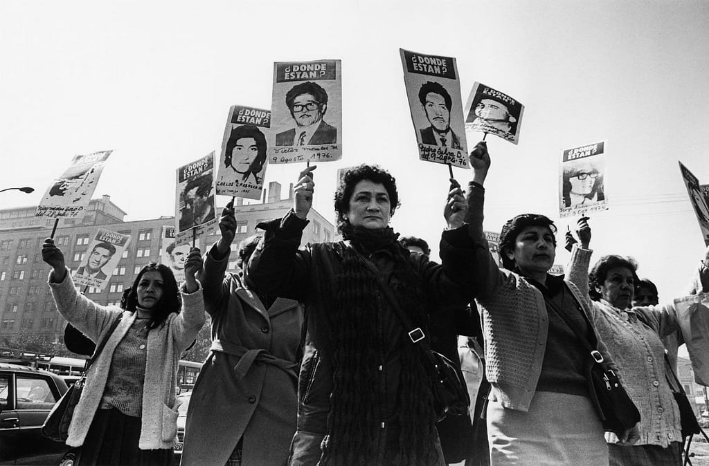 Chilean mothers of the “disappeared” gather, holding signs of their missing loved ones. (Photo by Kena Lorenzini, from Wikimedia Commons)