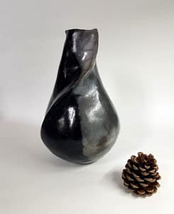 Druppelvorm met een schuine "vouw" van beneden naar boven. De linkerkant is diepzwart. De rechterkant grijs/zwart gewolkt. Voor de maat ligt er een dennenappeltje bij.